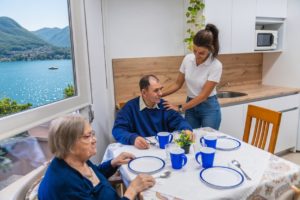 Operatrice domiciliare sorride e assiste una coppia di anziani in terrazza