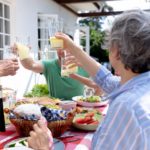 Gruppo di anziani che brindano con bicchieri di succo di frutta durante un pasto all'aperto su una terrazza soleggiata.
