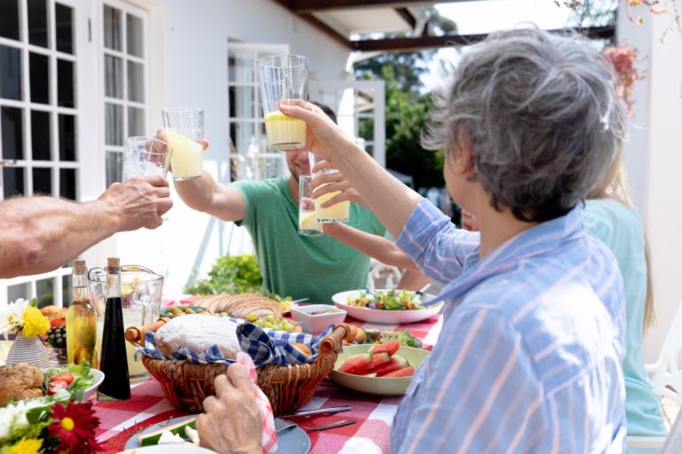 Gruppo di anziani che brindano con bicchieri di succo di frutta durante un pasto all'aperto su una terrazza soleggiata.