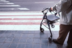 Anziano con deambulatore fermo vicino alle strisce pedonali, simbolo della difficoltà a camminare e del bisogno di sostegno nella mobilità.