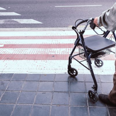 Anziano con deambulatore fermo vicino alle strisce pedonali, simbolo della difficoltà a camminare e del bisogno di sostegno nella mobilità.
