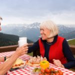 Coppia di anziani che condivide un pasto all'aperto su una terrazza panoramica con vista sulle Alpi svizzere.